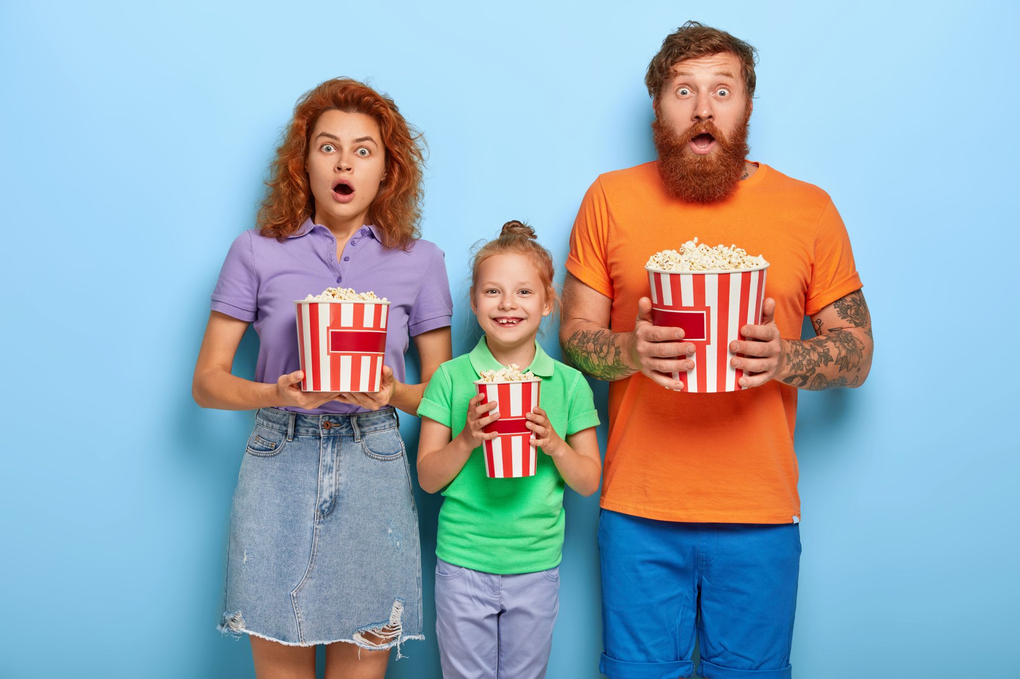 Three family members enjoy watching television at home, mother and father stare with frightened expressions at screen, their small daughter stands between and smiles, eating popcorn.