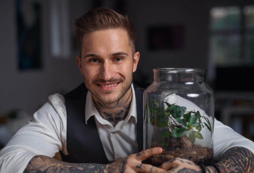 A tattooed businessman with plant terrarium sitting at the desk indoors in office.