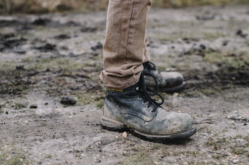 A muddy pair of black work boots worn with brown trousers by an unknown person in a dirty field