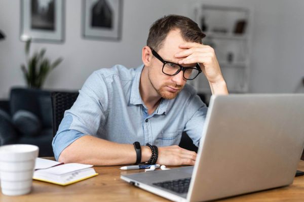 Thoughtful young man in modern glasses and casual wear looking on laptop screen. 