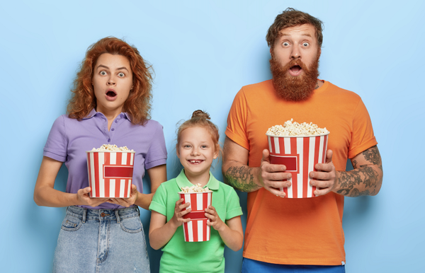 Mother and father stare with frightened expressions, their small daughter stands between and smiles, eating popcorn.